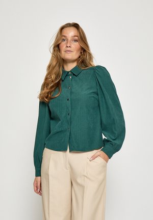 Türkisfarbenes Cordhemd mit Kragen, Knopfverschluss und aufgepufften langen Ärmeln, kombiniert mit beigen Hosen. Glatte Textur und dezente Rippung.