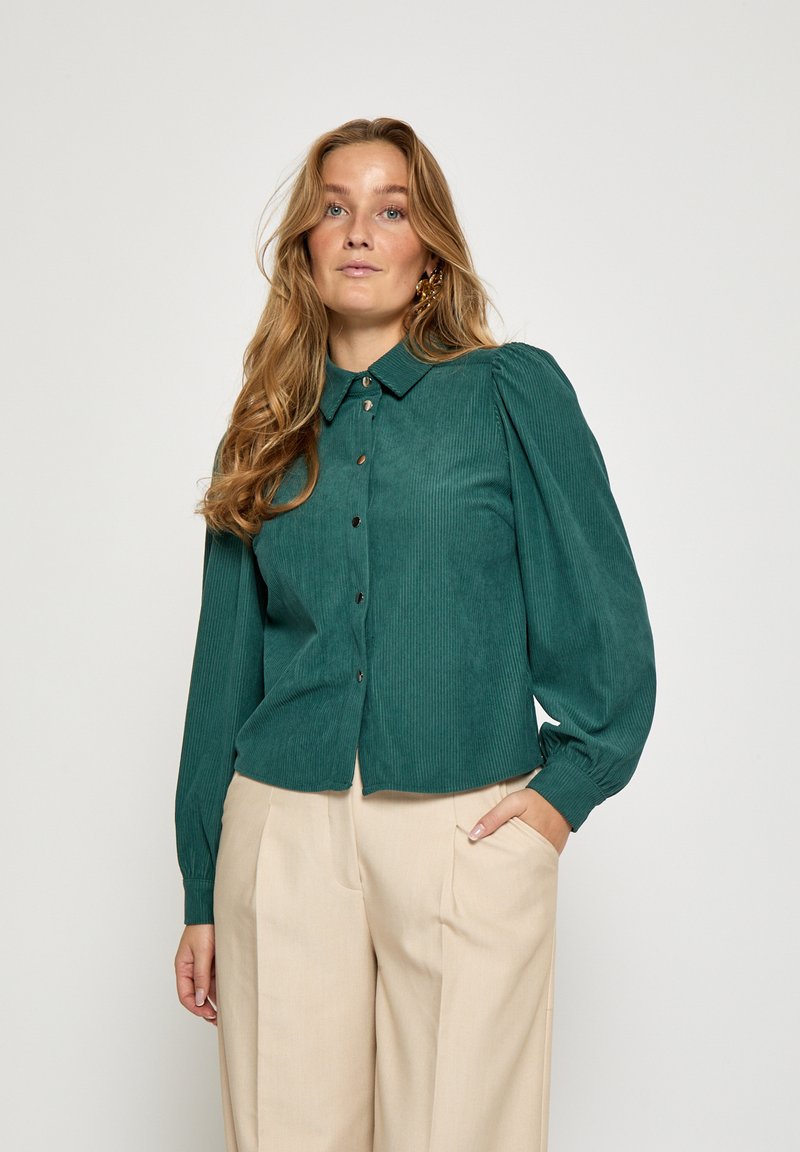 Žalsvos spalvos kordurojos marškiniai su apykakle, sagomis užsegamas priekyje ir pūstomis ilgomis rankovėmis, derinami su smėlio spalvos kelnėmis. Lygus audinys su subtiliomis gyslelėmis.