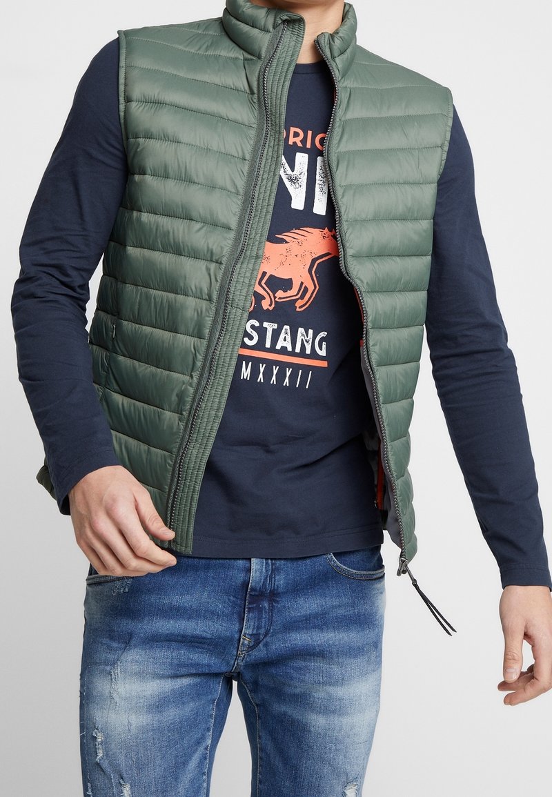 Grön, quiltad pufferväst med hög krage och dragkedja. Bärs över en marinblå långärmad skjorta med grafisk design. Jeans synliga nedan.