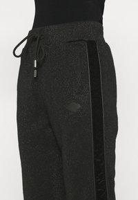 Schwarze Jogging-Hosen aus strukturiertem Material, mit einem Zugbandbund, Seitenstreifen und einem Rautenmuster-Logo-Patch auf dem Oberschenkel.