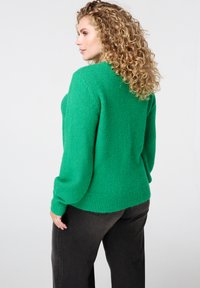 Groen gebreide trui met een losse pasvorm, geribbelde zoom en lange mouwen. De stof heeft een zachte textuur. Draagt bij zwarte wijde broekspijpen.