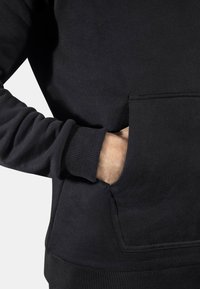 Sudadera negra con puños y dobladillo de canalé, que presenta un bolsillo frontal tipo canguro. Textura de tela suave, diseño minimalista, primer plano de una mano en el bolsillo.