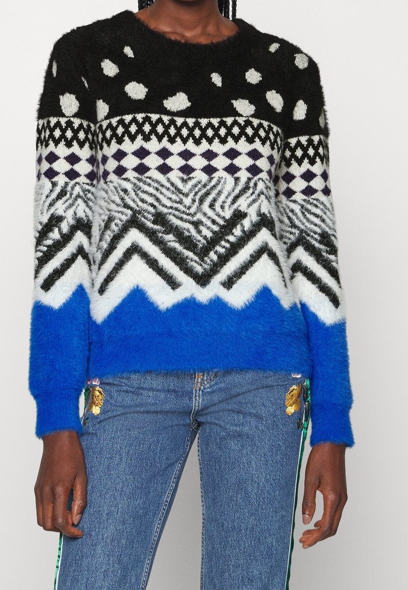 Femme portant un pull douillet avec des motifs géométriques et animaliers noirs, blancs et bleus, associé à un jean bleu avec broderies florales.