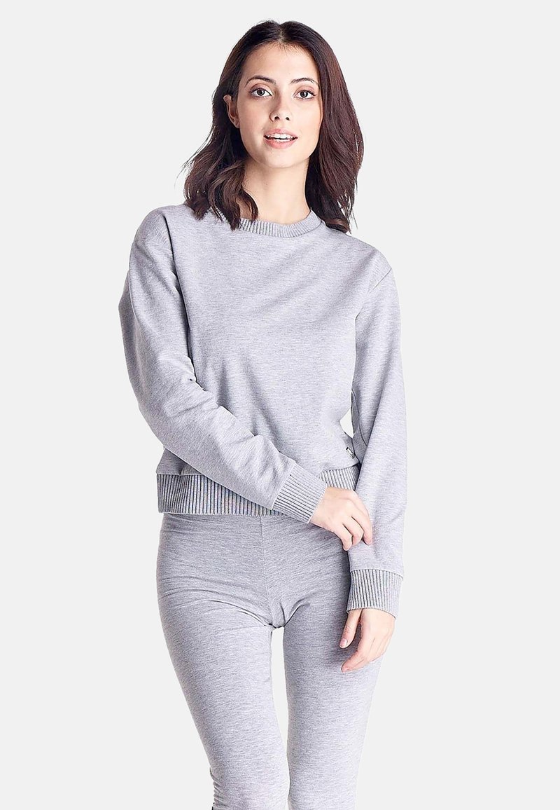 Jeune femme aux cheveux foncés mi-longs, portant un sweat-shirt gris à manches longues et un pantalon moulant assorti, sur fond blanc.
