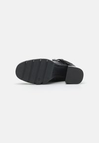 Zapato negro de tacón con una suela de goma sólida, que presenta un patrón texturizado y una correa decorativa con hebilla en la sección superior.