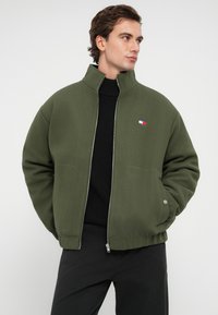 Ελαιόχρωμο αδιάβροχο μπουφάν με υψηλό γιακά, διαθέτει πλαϊνές τσέπες και μια μικρή ραφή λογότυπου. Φοριέται πάνω από ένα μαύρο κούμπωμα που έχει υφή.