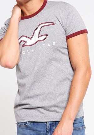 Personne portant un t-shirt gris Hollister avec des bordures et un logo bordeaux, associé à un jean bleu, la main touchant le cou sur un fond blanc.