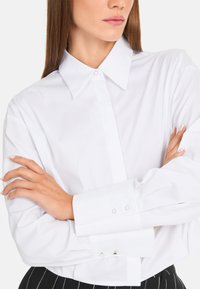 White long-sleeve shirt with a pointed collar, single button closure, and squared cuff details. Smooth fabric with a crisp appearance.