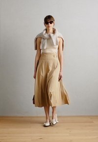 Jupe longue beige au design évasé, assortie à un haut ajusté. Un pull léger gris clair drape sur les épaules, complété par des chaussures blanches.