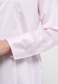 Camicia button-up rosa chiaro con maniche lunghe, caratterizzata da una texture liscia e un singolo bottone sui polsini. Design su misura con linee pulite.