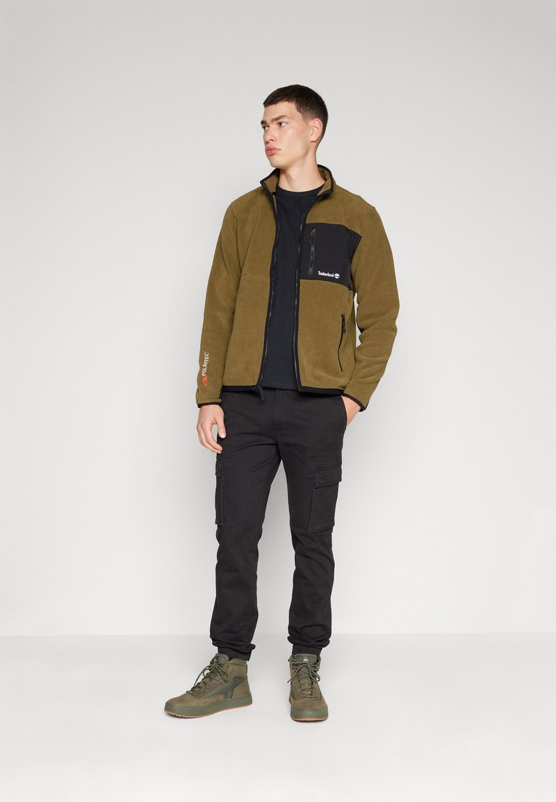 Kurtka polarowa w oliwkowym kolorze z czarną kieszenią, zamkiem błyskawicznym i wyhaftowanym logo. Połączona z czarnymi spodniami cargo i zielonymi butami.