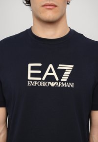 Temno modra bombažna majica s pomočjo kremne barve natisnjenim logotipom, ki vsebuje "EA7" in "EMPORIO ARMANI." Klasičen kroj s krožnim ovratnikom in kratkimi rokavi.