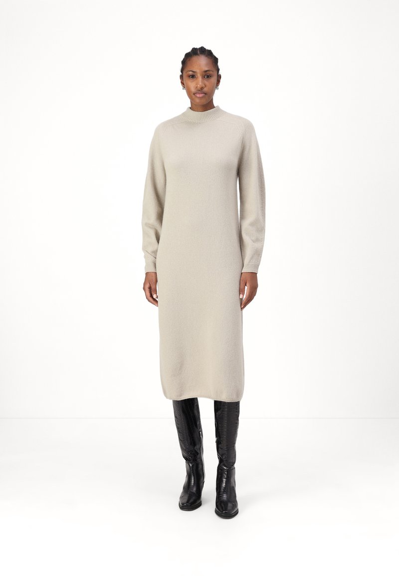 Robe tricotée beige à manches longues avec un col haut, texture lisse et silhouette droite, assortie à des bottes en cuir noires jusqu'aux genoux.