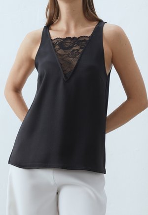 Kobieta w czarnym, bezrękawnym topie z koronkowym detalem na dekolcie w kształcie litery V, w połączeniu z białymi spodniami, stojąca na jasnym, jednolitym tle.