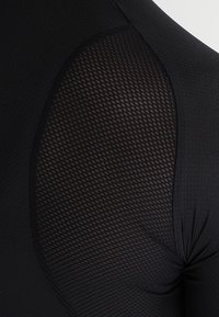 Top deportivo negro con un panel de malla texturizada debajo del brazo, tela suave en el cuerpo y detalle fruncido en la costura del hombro.