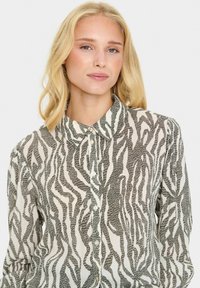 Žena s blond vlasmi, oblečená v košeli s dlhými rukávmi a gombíkmi s čiernobielym abstraktným vzorom zebry, stojí čelom proti jednoduchému pozadiu.