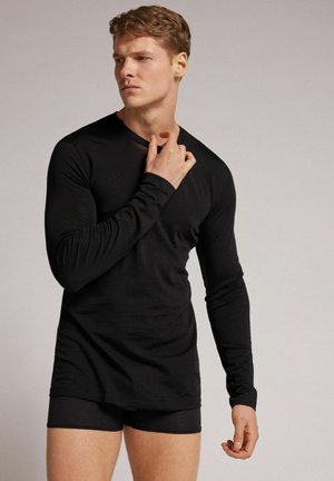 Junger Mann, der ein eng anliegendes schwarzes Langarmshirt und schwarze Shorts trägt, schaut seitlich mit einer Hand am Hals.