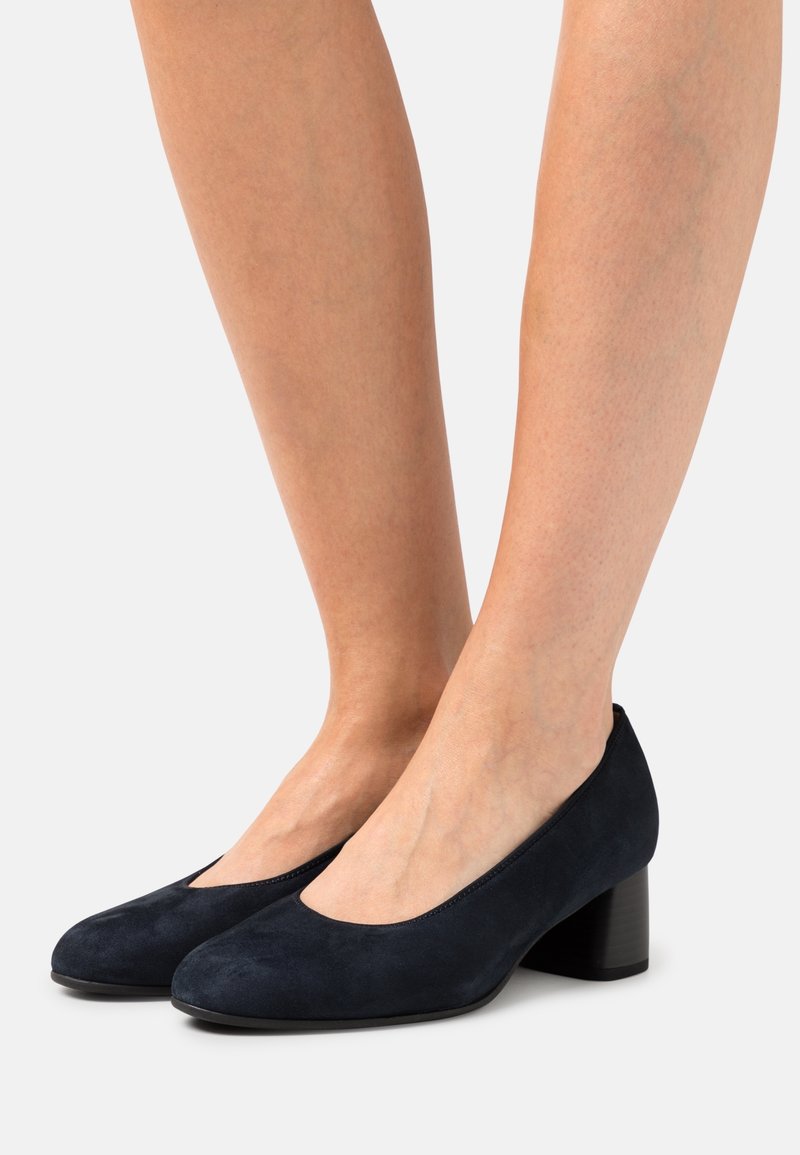 Femme portant des chaussures en daim noir à petit talon bloc et bouts arrondis, debout sur fond blanc.