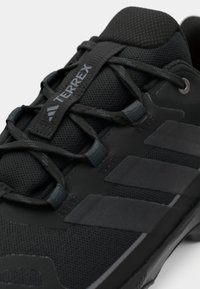 Schwarzer Sportschuh mit einem strukturierten Mesh-Obermaterial, kontrastierenden Gummielementen und drei markanten Streifen an der Seite.