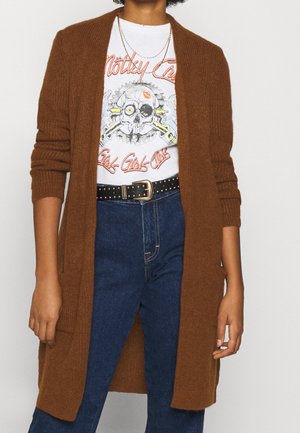 Cardigan en tricot marron avec un décolleté ouvert et des manches longues, porté sur un t-shirt à motifs. Jean bleu foncé avec une ceinture noire à clous.