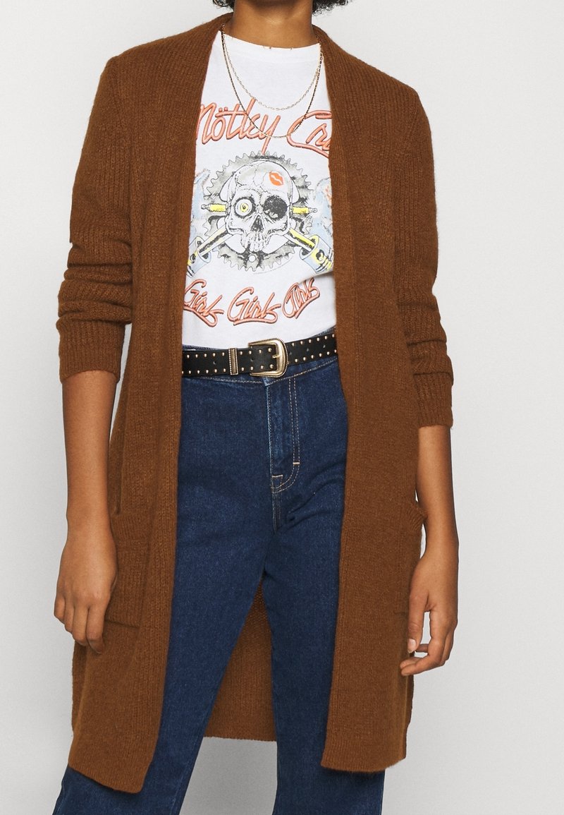 Brun stickad cardigan med öppen framsidan och långa ärmar, bärs över en grafisk t-shirt. Mörkblå jeans med ett svart nitted bälte.