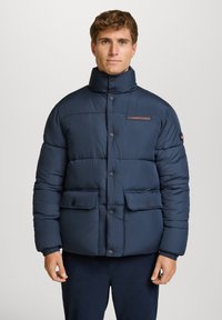 Giacca puffer blu navy con colletto alto, bottoni frontali, due tasche grandi e etichetta con marchio sul petto. Design texturizzato e imbottito.