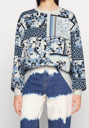 Kobieta ubrana w patchworkowy top w kwiaty i kratę w kolorach niebieskim, czarnym i białym z długim rękawem, zestawiony z dżinsami w stylu tie-dye w odcieniach niebieskiego i białego.