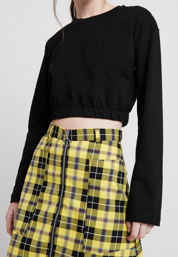 Femme portant un haut court à manches longues noir et une jupe à carreaux jaunes avec une fermeture éclair frontale et des poches.