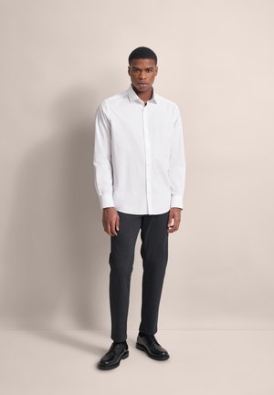 Mann steht vor einem beigen Hintergrund, trägt ein weißes Button-up-Hemd, dunkelblaue Slim-Fit-Hosen und schwarze Dressschuhe.