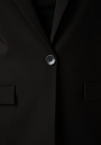 Close-up van een zwarte blazer, dichtgeknoopt in het midden, met aan elke kant gedeeltelijk zichtbare klepzakken.