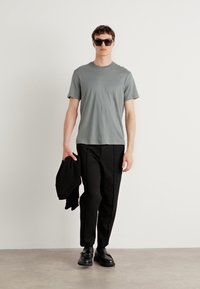 Tricou din bumbac gri, pantaloni negri, pantofi negri și ochelari de soare. Modelul ține un sacou negru. Design simplu cu o croială lejeră.