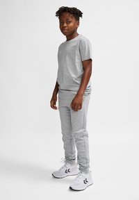 Graues Baumwoll-T-Shirt und Jogginghose, beide mit glatter Textur. Weiße Sneaker mit schwarzen Akzenten. Stehende Pose mit leichtem Seitenblick.