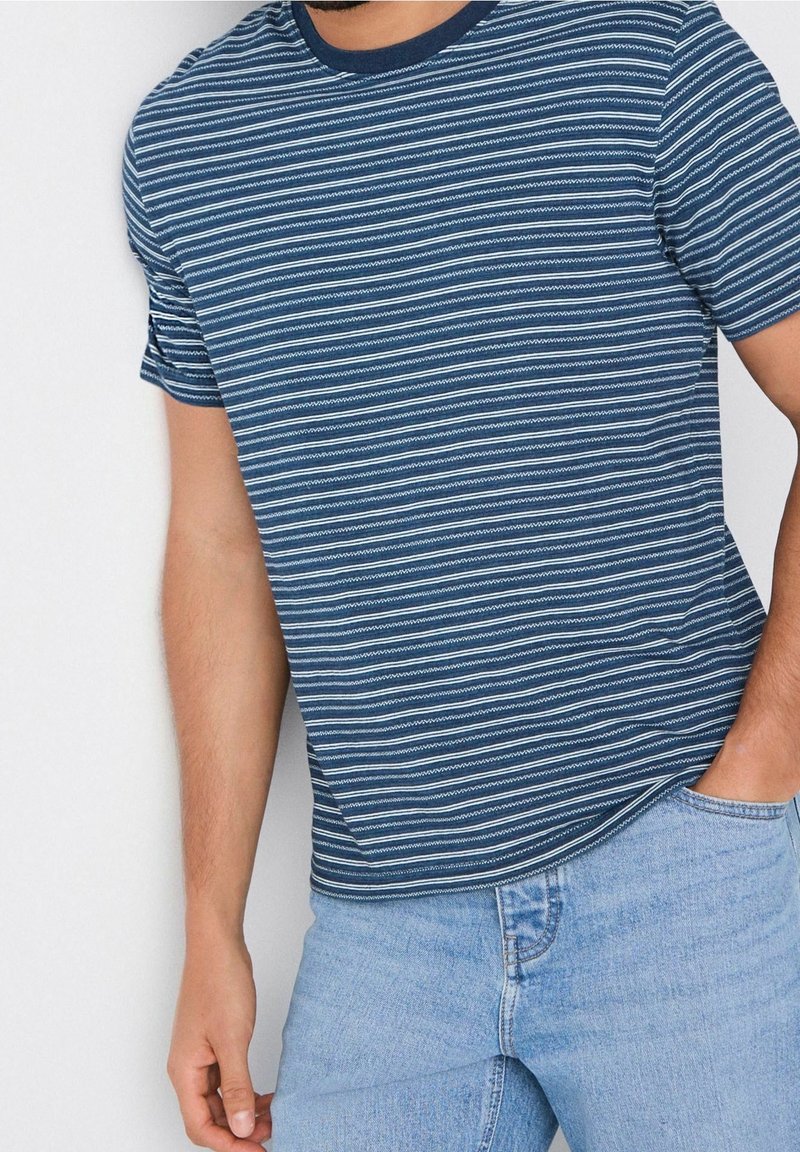 Homme portant une chemise à manches courtes rayée bleu et blanc et un jean bleu clair, debout avec une main dans la poche.