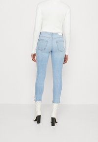 Los skinny jeans azul claro presentan una cintura alta, bolsillos traseros y aberturas en el dobladillo, combinados con una blusa blanca de manga larga y botas de tobillo color crema.