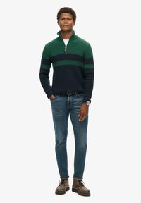 Sweter zapinany na zamek w ciemnym granacie, z zielonymi i granatowymi paskami. Połączony z jasnoniebieskimi dżinsami i brązowymi butami na sznurówki.