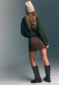 Pull vert foncé, mini-jupe marron et bottes noires montantes avec plusieurs boucles. Les longs cheveux du mannequin tombent en cascade dans son dos.