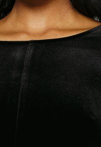 Zwarte fluwelen top met een brede halslijn, voorzien van een centrale naad en een soepele textuur. De stof weerkaatst subtiel licht, wat de diepte ervan versterkt.