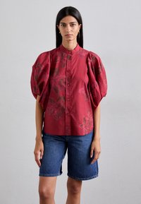 Rote Bluse mit Blumenmuster, gepufften Ärmeln, Knopfleiste und weicher Textur. Kombiniert mit Denim-Bermuda-Shorts. Schlichtes, modernes Design.