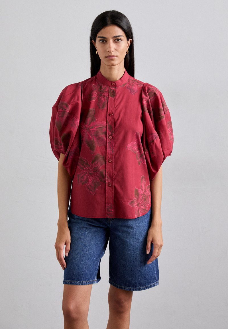 Red floral blouse with puffed sleeves, button front, and a soft texture. Paired with denim Bermuda shorts. Simple, modern design.