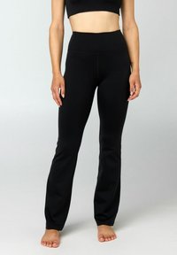 Vrouw die zwarte, hoog getailleerde, nauwsluitende broek en een zwart cropped top draagt, blootsvoets staand tegen een effen lichte achtergrond.