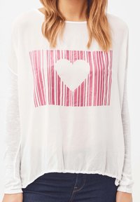 Top blanco ligero con rayas verticales rosas y un gráfico de corazón central. Textura suave con mangas largas transparentes y un dobladillo redondeado.