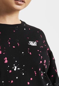 Mujer con sudadera negra con manchas de pintura rosa y blanca y logotipo estilizado bordado en blanco en la parte superior del pecho.