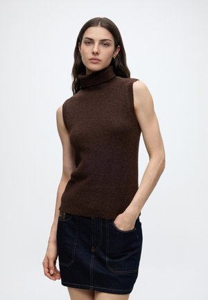Kvinde med langt mørkt hår iført brun ærmeløs rullekrave og mørkt denimskørt, står med den ene hånd i lommen mod en ensfarvet baggrund.