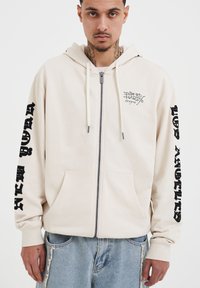Felpa beige con zip, con lettering nero sulle maniche, logo sul davanti, tasca a marsupio e cappuccio con cordino. Materiale morbido e vestibilità rilassata.