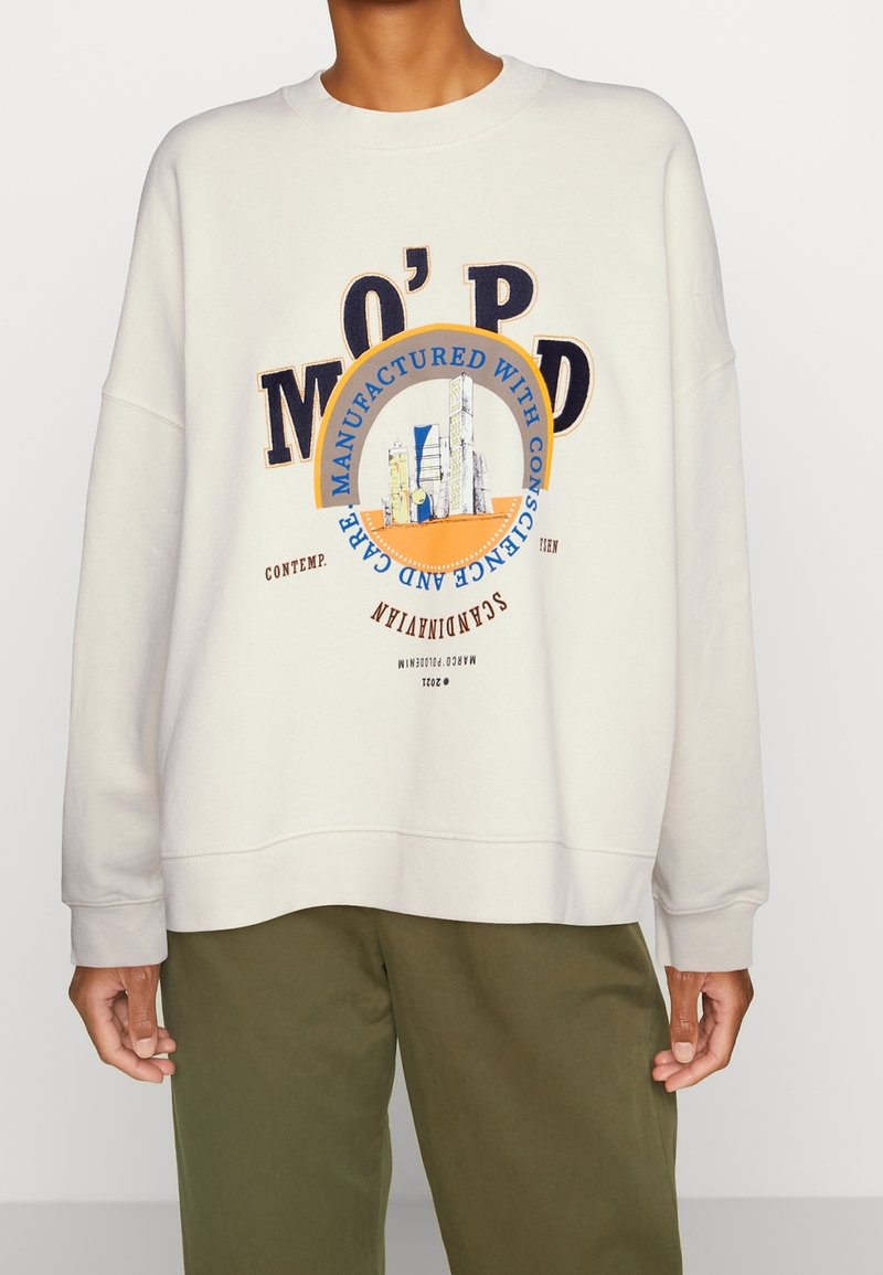 Persona vistiendo un sudadera blanco sucio con un gráfico de un horizonte urbano y texto, combinado con pantalones verde oliva, de pie contra un fondo plano.
