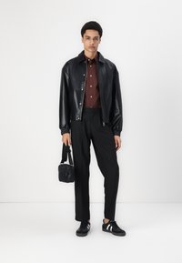 Giacca di pelle nera con polsini elastici, camicia bordeaux con bottoni, pantaloni neri e sneakers nere con strisce bianche. Tiene una piccola borsa nera.