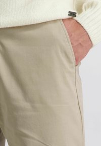 Hand in de zak van beige broek gedragen met een witte geribbelde trui met een klein zwart label.