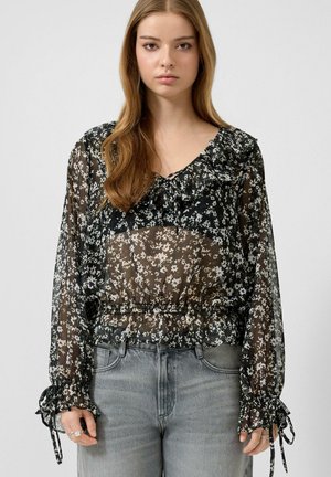 Młoda kobieta z długimi włosami, ubrana w przezroczystą czarną kwiecistą bluzkę z falbanami i zawiązywanymi mankietami, w połączeniu z jasnoszarymi jeansami.