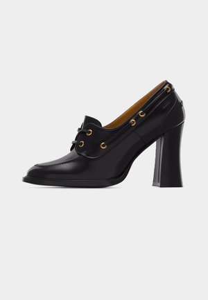 Musta nahkainen korkokengällä varustettu loafer, jossa on kultaiset silmukat, nyöritys, pyöreä kärki ja tilava lohkokorko, yksinkertaisella valkoisella taustalla.