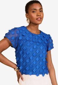 Top trasparente blu con applicazioni floreali 3D, maniche corte, scollatura tonda e bordo decorativo. Tessuto leggero e dalla texture liscia.